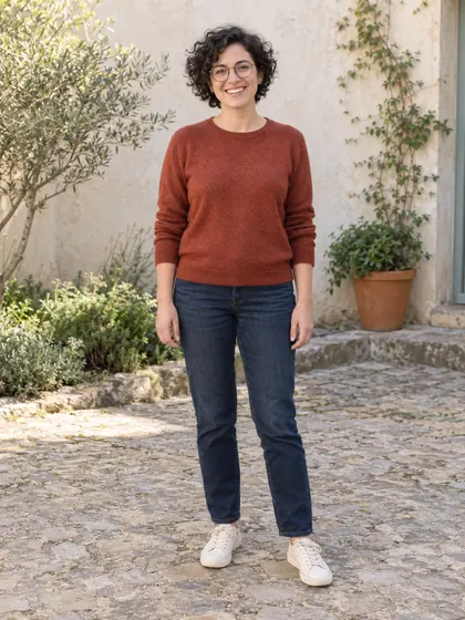 AI-generated photo of a fictional adult with curly hair and round glasses, wearing a rust-red sweater in a leafy courtyard